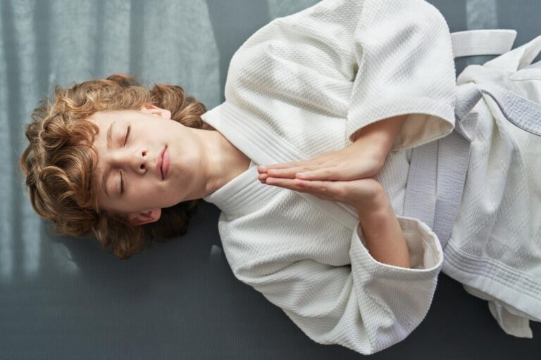 top-view-curly-haired-preteen-kid-kimono-lying-floor-with-closed-eyes-hands-namaste-gesture-judo-training_935394-6175